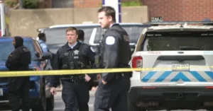 Police officers are seen at a crime scene with a yellow tape barrier and a police vehicle in the background.
