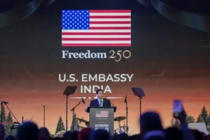 The image shows a person speaking at a podium at the U.S. Embassy in India, celebrating the 250th anniversary of American independence.