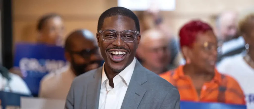 A man in a suit smiles brightly in front of a group of people at what appears to be a public event or gathering.