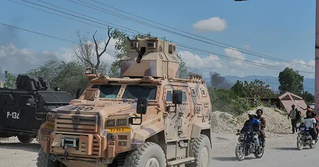 The image shows a military armored vehicle parked on a road, with people on motorcycles passing by in a rural setting.