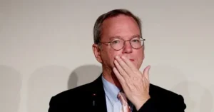 The image shows a man in a suit and tie, wearing glasses, with his hand near his mouth, possibly in a thoughtful or contemplative pose.