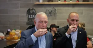 Two men in suits are drinking from small cups in a casual setting, with a brick wall and a "RIP" sign in the background.