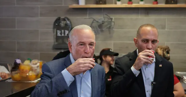 Two men in suits are drinking from small cups in a casual setting, with a brick wall and a "RIP" sign in the background.