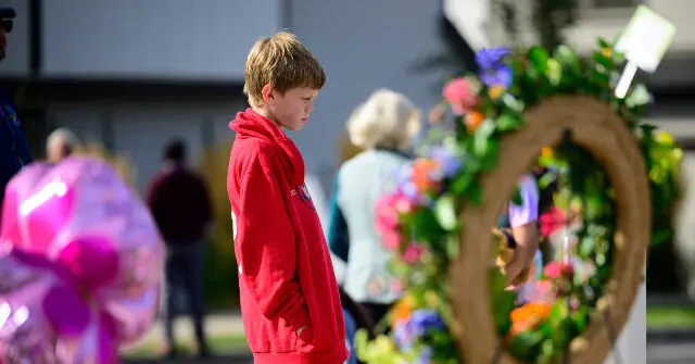 A young boy in a red hoodie stands solemnly in front of a floral memorial during a community event.