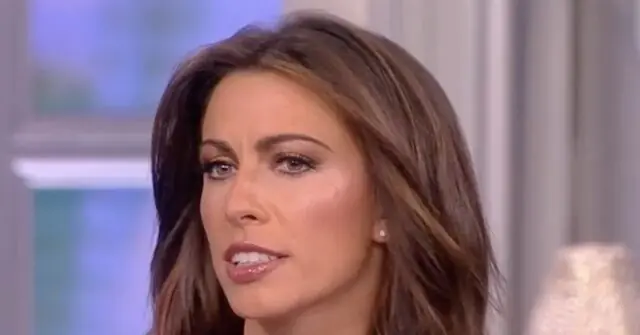 The image shows a person with long, wavy hair, wearing a light-colored top, in what appears to be a television studio setting.