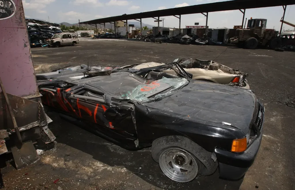 The image shows a damaged and partially dismantled car in a junkyard, surrounded by other wrecked vehicles.