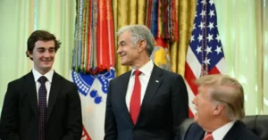 The image shows three individuals in formal attire, with one seated and two standing, in a setting that includes a U.S. flag and other national flags in the background.