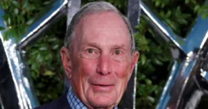 The image shows a man in a suit standing in front of a backdrop with a metallic, reflective design.