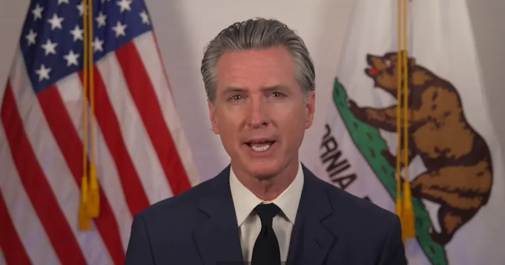 The image shows a man in a suit speaking in front of the American and California state flags.