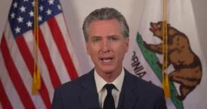 The image shows a man in a suit speaking in front of the American and California state flags.