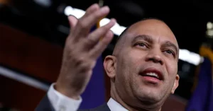 The image shows a man in a suit gesturing with his hand, appearing to speak or present in a formal setting.