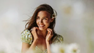 A woman in a floral dress smiles brightly, holding her hands near her face, with a flower in her hair, set against a soft, blurred background.