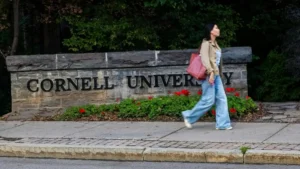A person walks past a stone sign reading "Cornell University" on a campus pathway.