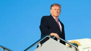 The image shows a person in a suit standing on the steps of an aircraft, with a clear blue sky in the background.