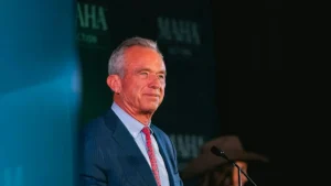 The image shows a man in a suit and tie standing at a podium, likely during a formal event or speech.