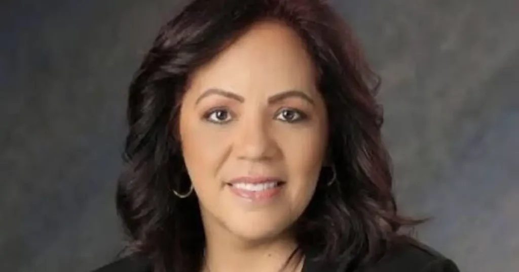 The image shows a woman with dark, wavy hair, wearing a black top and hoop earrings, against a neutral background.