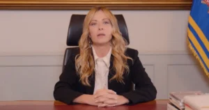 A woman in a professional setting, seated at a desk with a flag in the background, appears to be in a formal or official capacity.