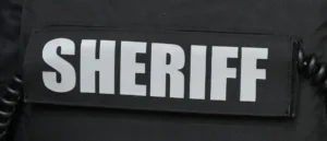 The image shows a close-up of a black uniform with a white "SHERIFF" patch.