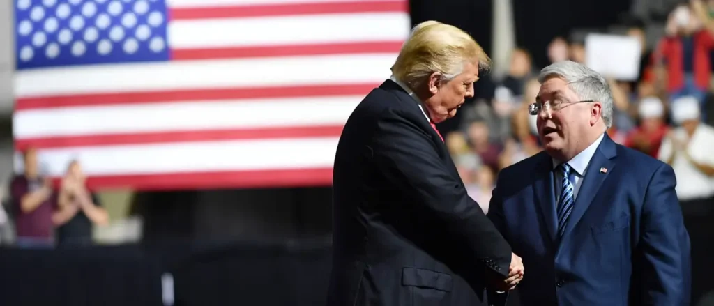 Two men in suits shake hands in front of an American flag, with a crowd of people in the background.