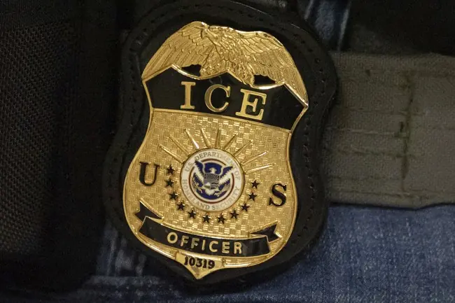 The image shows a close-up of a U.S. Immigration and Customs Enforcement (ICE) officer's badge.