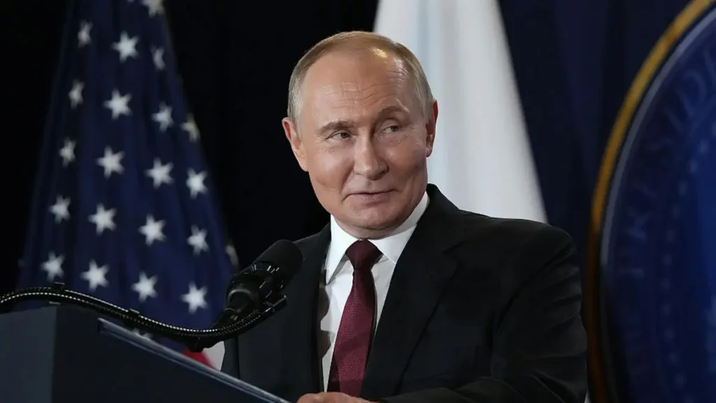 The image shows a man in a suit standing at a podium with a microphone, with flags of the United States and another country in the background.
