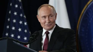 The image shows a man in a suit standing at a podium with a microphone, with flags of the United States and another country in the background.