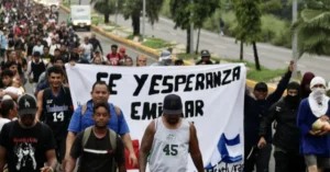 A group of people march down a street holding a banner that reads "FE Y ESPERANZA EMIGRAR," expressing hope and faith amidst migration.