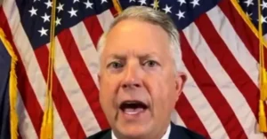 The image shows a person speaking in front of multiple American flags.