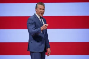 A man in a suit gestures while speaking in front of a backdrop featuring red and white stripes.