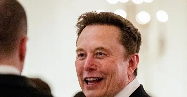 The image shows a man in a suit smiling and engaged in conversation with another individual.