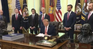 The image shows a formal setting with a seated individual at a desk, surrounded by several men in suits, with American flags and other official decorations in the background.