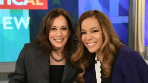 Two women are smiling and posing together on a news set, with a colorful background and the word "NEW" visible.