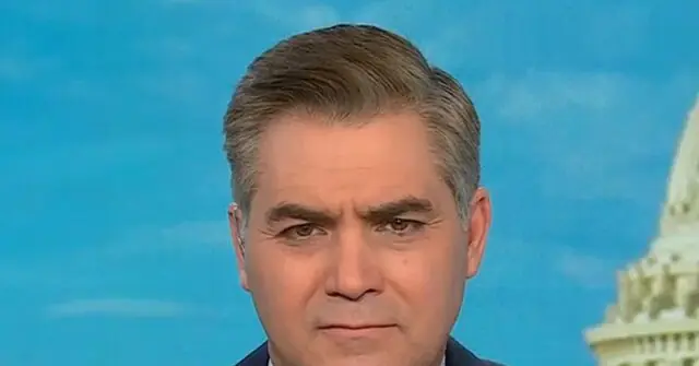 The image shows a news anchor in a studio setting with a backdrop of a clear blue sky and a partial view of a building resembling the U.S. Capitol.