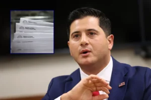 The image shows a man in a suit speaking, with a smaller inset image of what appears to be voter registration forms.