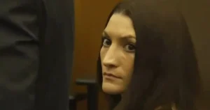 The image shows a person with long dark hair, wearing a dark garment, in what appears to be a courtroom setting.