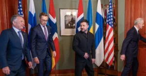 The photo shows a group of men in formal attire standing in front of various national flags, likely during a diplomatic or political meeting.