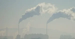 The image shows industrial smokestacks emitting thick plumes of smoke into the air, indicating significant air pollution.