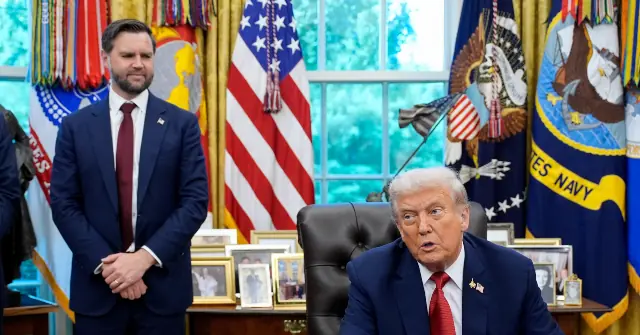The image depicts a formal setting with two individuals in suits, one seated at a desk and the other standing, in front of a backdrop featuring American flags and military insignia.