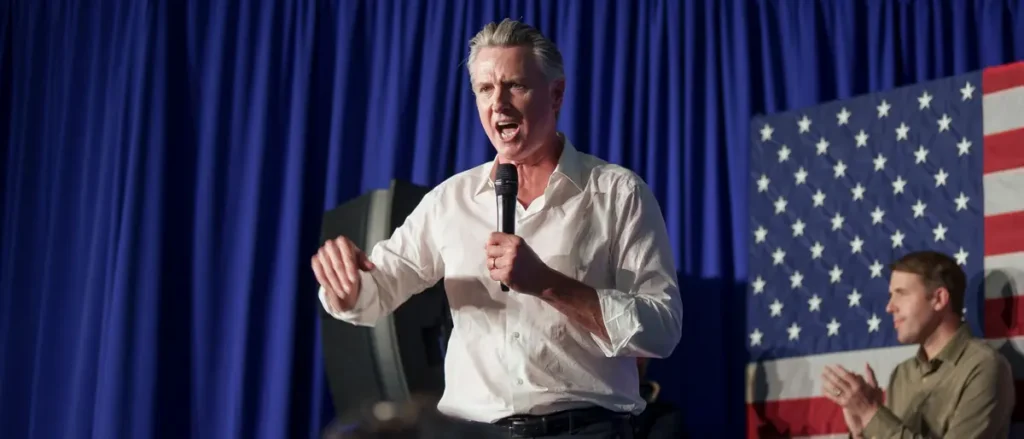A man is speaking passionately into a microphone at a political rally, with an American flag in the background.