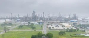 The image shows an industrial complex with numerous tall structures and smokestacks, likely an oil refinery, under a cloudy sky.