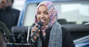 A woman wearing a patterned hijab and scarf speaks into a microphone in front of a vehicle.