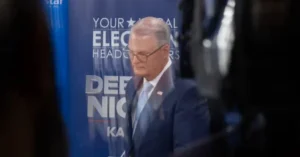 The image shows a man in a suit standing in front of a backdrop with political campaign text.