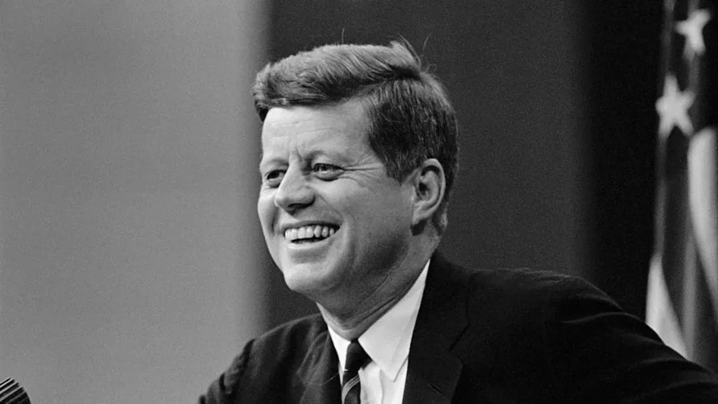The image shows a smiling man in a suit, with an American flag in the background, suggesting a formal or political setting.