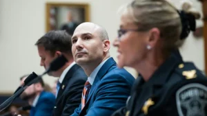 A man in a suit and a woman in a police uniform are seated at a table with microphones, likely participating in a formal meeting or hearing.