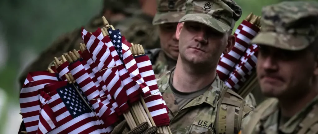 A group of soldiers in camouflage uniforms carry American flags during a solemn ceremony.