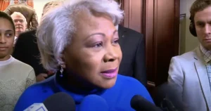 A woman in a blue turtleneck is speaking to reporters surrounded by microphones and other individuals.