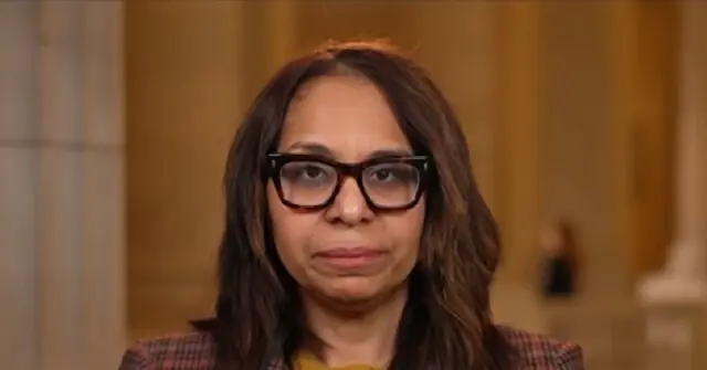 The image shows a person wearing glasses and a plaid blazer, set against a blurred background that appears to be an indoor setting with columns.