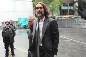 A man in a suit walks through a city street, with a police officer and another individual in the background.