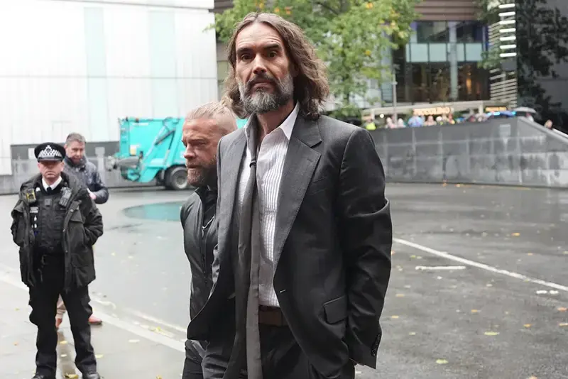 A man in a suit walks through a city street, with a police officer and another individual in the background.