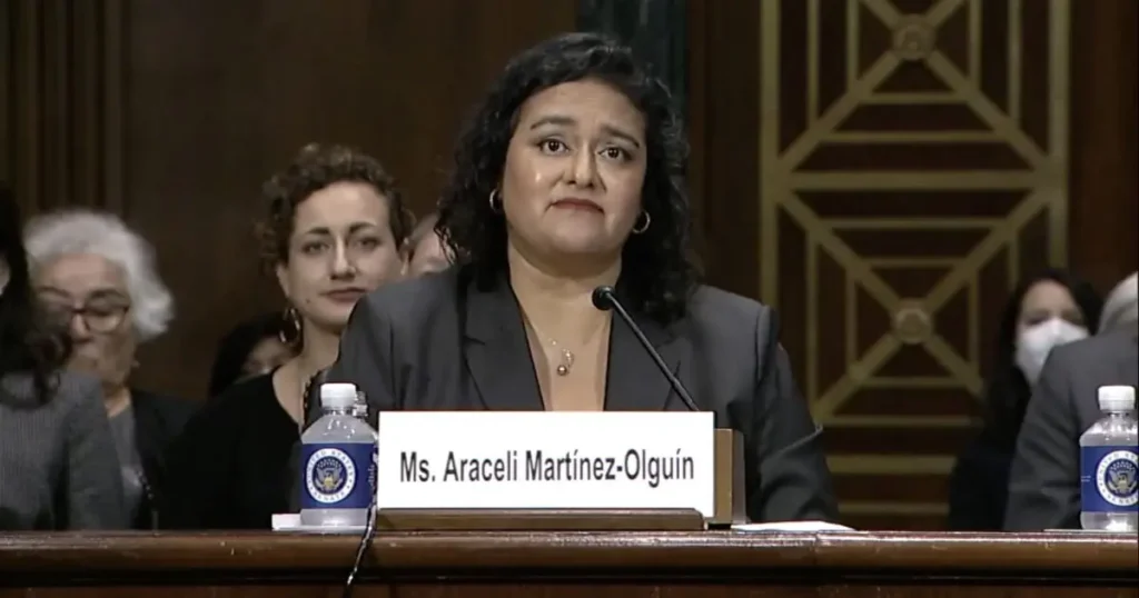 A woman is seated at a table with a nameplate that reads "Ms. Araceli Martínez-Olguín," suggesting she is participating in a formal hearing or meeting.